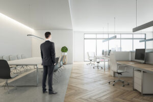 Pensive young businessman in suit standing in wooden coworking meeting room interior with daylight, furniture and equipment. Design and ceo worker concept.